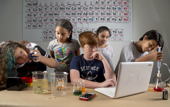 Students in front of a periodic table of elements using scientific equipment and technology.
